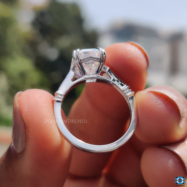 Milgrain ring featuring emerald cut moissanite and round side stone accents.