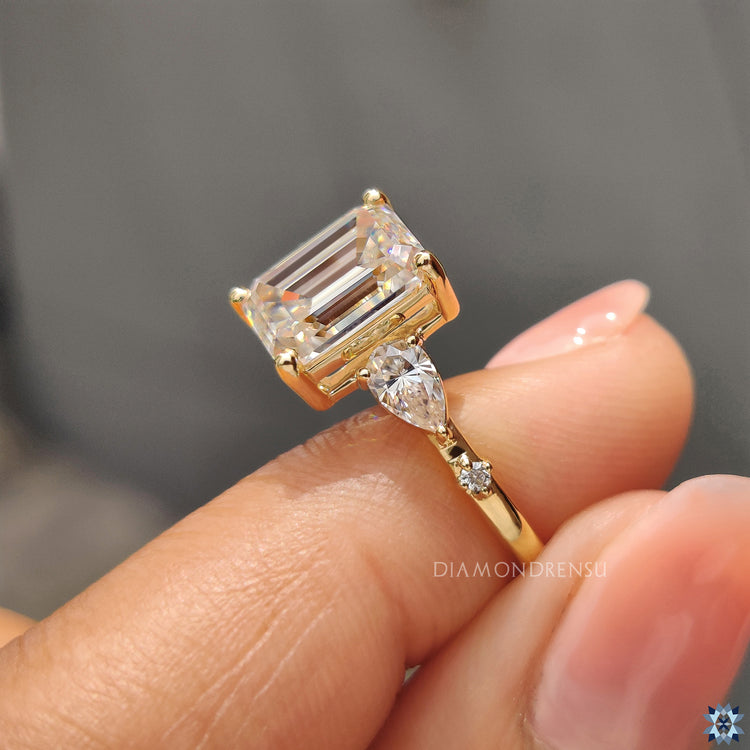 Side stone moissanite engagement ring in yellow gold with emerald cut centre.