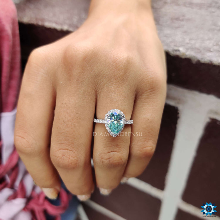Moissanite halo ring featuring a cyan blue pear cut stone in prong setting.