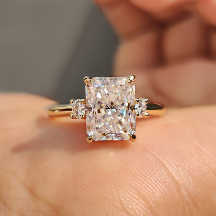 Three stone ring featuring radiant cut moissanite and round side stones.
