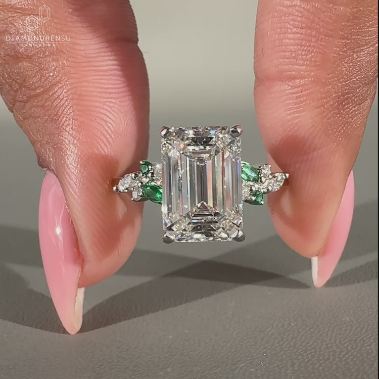 White gold engagement ring with green emerald side stones and prong setting.
