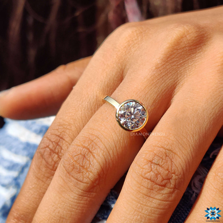 Round cut Moissanite engagement ring with bezel design and handmade yellow gold band.

